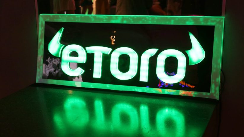 eToro logo in bright green neon light, reflecting on a dark, glossy surface.
