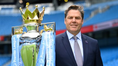 Richard Masters, Premier League CEO, smiling next to the Premier League trophy with stadium seats in background