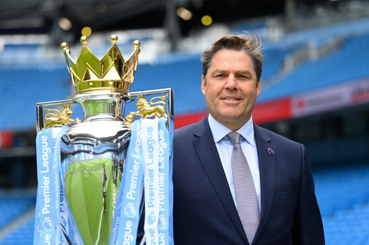 Richard Masters, Premier League CEO, smiling next to the Premier League trophy with stadium seats in background