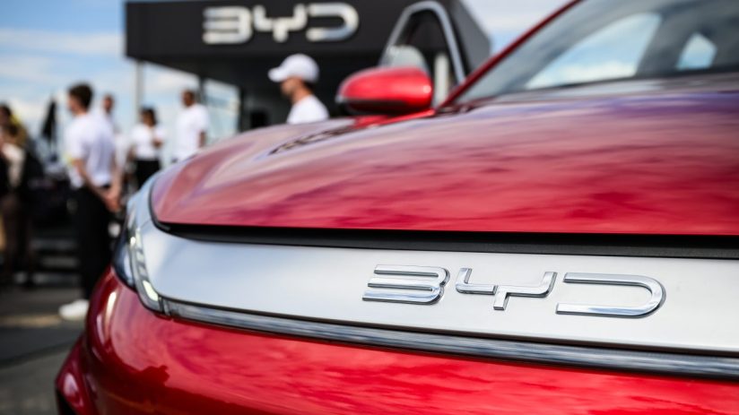 Close-up of a red BYD car with the brand logo prominent on the front, blurred figures and a BYD sign in the background.