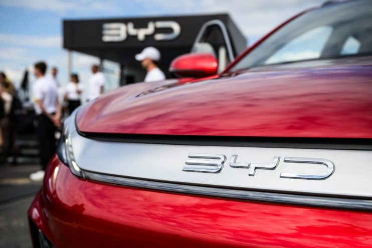 Close-up of a red BYD car with the brand logo prominent on the front, blurred figures and a BYD sign in the background.