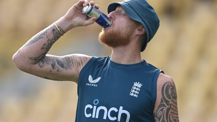 Ben Stokes, with a red beard and bucket hat, drinks from a Red Bull can while wearing an England cricket vest.