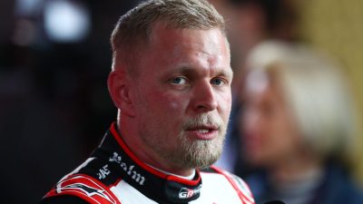 Man with blond hair and beard wearing a racing suit, speaking into a microphone at a news event.
