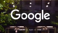 Illuminated white Google logo on a dark building facade with green tree reflections and a revolving door.