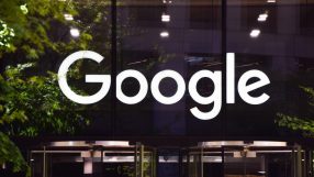 Illuminated white Google logo on a dark building facade with green tree reflections and a revolving door.