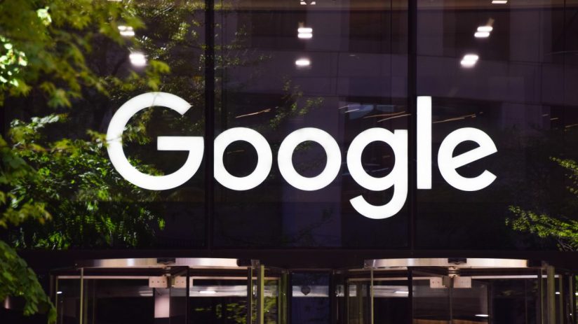 Illuminated white Google logo on a dark building facade with green tree reflections and a revolving door.