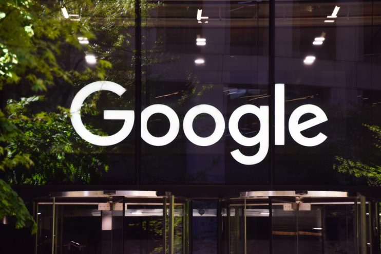Illuminated white Google logo on a dark building facade with green tree reflections and a revolving door.