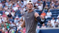 Tennis player in a gray sleeveless shirt and white cap celebrates with a clenched fist on court