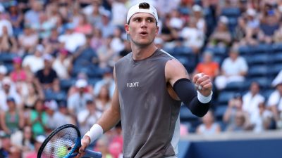 Tennis player in a gray sleeveless shirt and white cap celebrates with a clenched fist on court