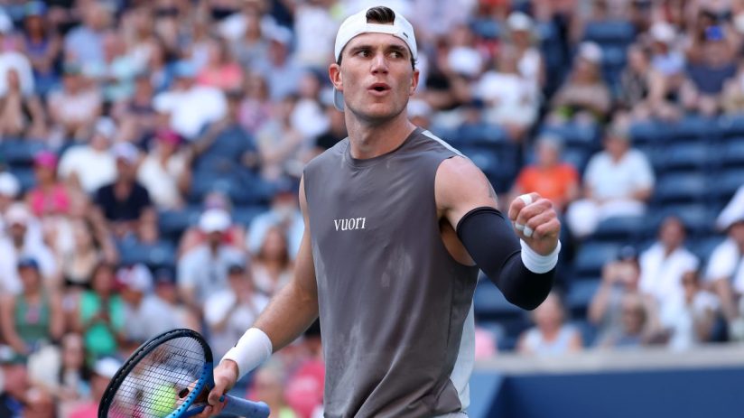 Tennis player in a gray sleeveless shirt and white cap celebrates with a clenched fist on court