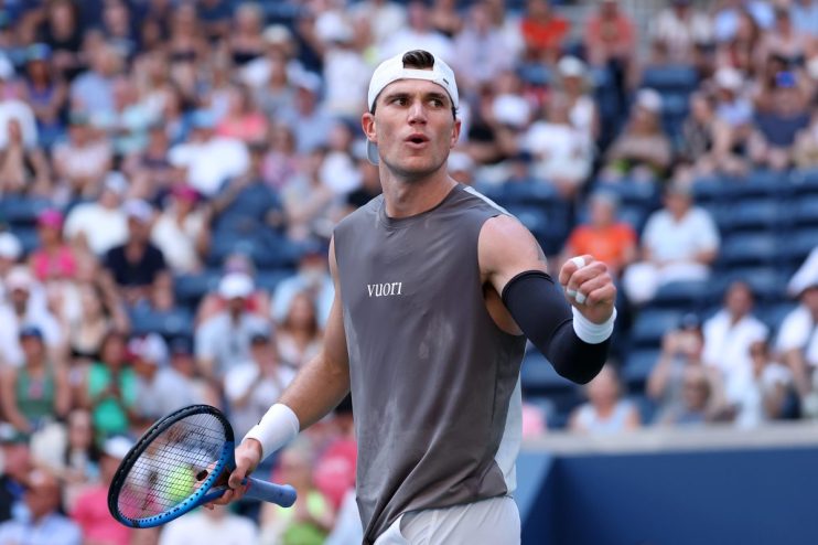 Tennis player in a gray sleeveless shirt and white cap celebrates with a clenched fist on court