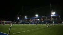 Rugby players warming up on a floodlit stadium field at night, Peter Thomas Stand visible in background.