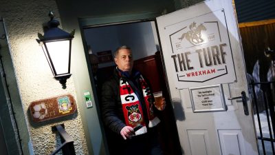 A man in a Wrexham AFC scarf holds a pint of beer, exiting The Turf pub with a Closed Until 12:00 sign.