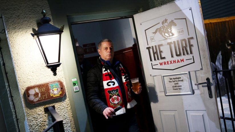 A man in a Wrexham AFC scarf holds a pint of beer, exiting The Turf pub with a Closed Until 12:00 sign.