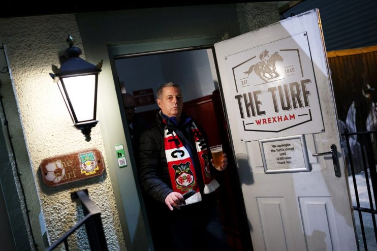 A man in a Wrexham AFC scarf holds a pint of beer, exiting The Turf pub with a Closed Until 12:00 sign.