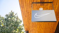 Nike swoosh logo sign hanging from a modern wooden building facade, with a green tree and blue sky in the background.