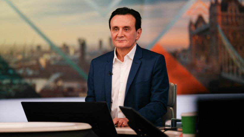 Man in blue suit and white shirt seated at a desk with blurred London cityscape and Tower Bridge background