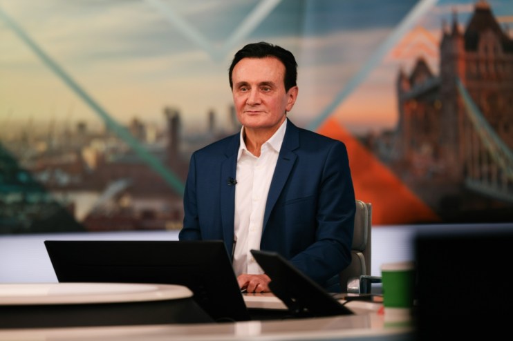 Man in blue suit and white shirt seated at a desk with blurred London cityscape and Tower Bridge background