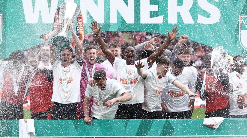 Football players celebrating their Vertu Trophy Final 2026 win under a Winners banner, champagne spraying