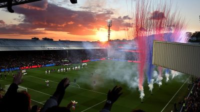 Football stadium at sunset with red and blue smoke flares, players on field, and fans in stands.