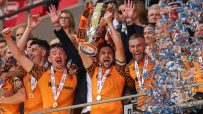 Jubilant football players in orange and black shirts celebrate with a trophy and medals as confetti falls.