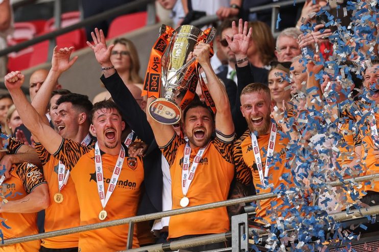 Jubilant football players in orange and black shirts celebrate with a trophy and medals as confetti falls.