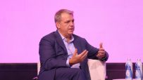 Businessman in a blue suit speaking on stage with hand gestures, two water bottles visible.