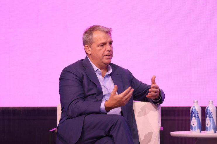 Businessman in a blue suit speaking on stage with hand gestures, two water bottles visible.