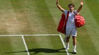 Tennis player waving to the crowd on a grass court, red bag over shoulder, white uniform