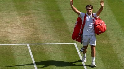 Tennis player waving to the crowd on a grass court, red bag over shoulder, white uniform
