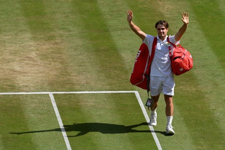 Tennis player waving to the crowd on a grass court, red bag over shoulder, white uniform