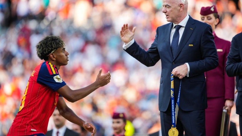 FIFA President Gianni Infantino with a gold medal, interacting with a young footballer in a red jersey.
