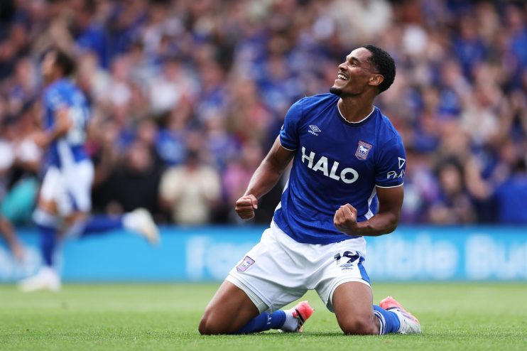 Footballer in a blue jersey celebrating on his knees on a green field with fans in the background.