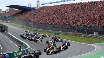 Formula 1 race cars on track with a packed grandstand of cheering fans at a major motorsport event.