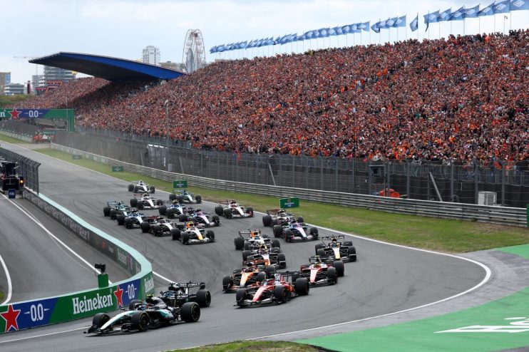 Formula 1 race cars on track with a packed grandstand of cheering fans at a major motorsport event.