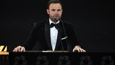 Juan Mata speaking at a podium with microphones, wearing a black velvet tuxedo and bow tie.