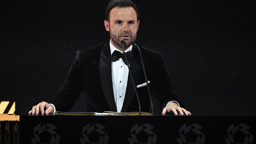 Juan Mata speaking at a podium with microphones, wearing a black velvet tuxedo and bow tie.