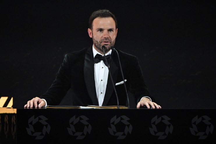 Juan Mata speaking at a podium with microphones, wearing a black velvet tuxedo and bow tie.