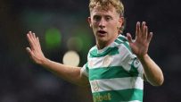 Blonde male football player in green and white striped Celtic jersey raising hands on field