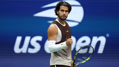 Tennis player in brown and white athletic wear, holding a racket, fist clenched, US Open background