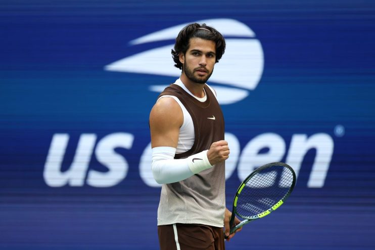 Tennis player in brown and white athletic wear, holding a racket, fist clenched, US Open background