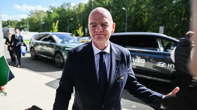 Gianni Infantino, FIFA President, in a navy suit and tie, speaking outdoors with cars and trees in the background.
