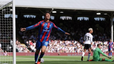 Footballer in red and blue jersey celebrating a goal with arms outstretched on a sunny field