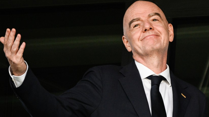 Gianni Infantino, FIFA President, in a dark suit and tie, smiling and raising his hand.
