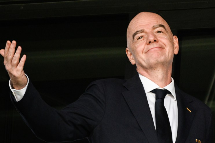 Gianni Infantino, FIFA President, in a dark suit and tie, smiling and raising his hand.