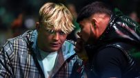 Two men, one with blonde hair and a plaid jacket, the other with dark hair and a puffer jacket, conversing closely.