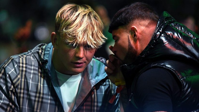 Two men, one with blonde hair and a plaid jacket, the other with dark hair and a puffer jacket, conversing closely.