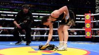 Female boxer Katie Taylor picks up her boxing gloves in the ring after a match, with a cameraman nearby.