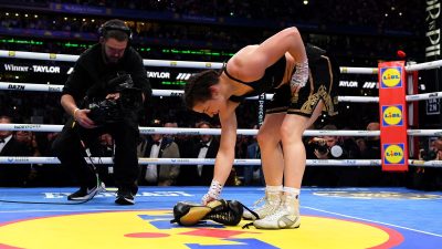 Female boxer Katie Taylor picks up her boxing gloves in the ring after a match, with a cameraman nearby.