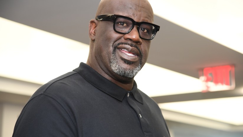 Shaquille ONeal in a black polo shirt and black-framed glasses, smiling slightly, with a blurred background.